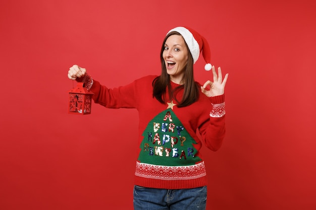 Foto alegre joven santa con sombrero de navidad que muestra un gesto de ok sosteniendo un candelabro de linterna vintage aislado sobre fondo rojo. feliz año nuevo 2019 celebración concepto de fiesta navideña. simulacros de espacio de copia.