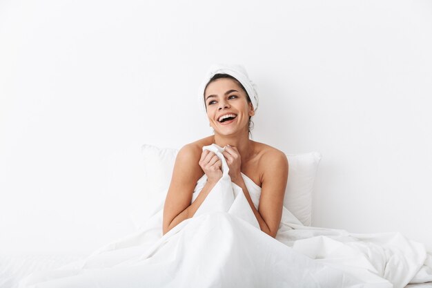 Alegre joven sentada en la cama después de la ducha envuelta en una manta, posando