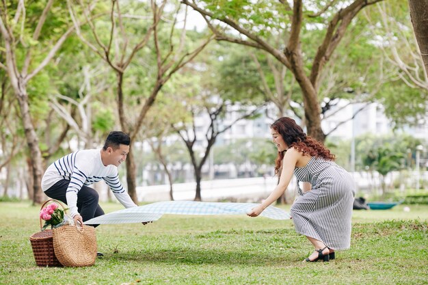 Alegre joven sonriente novio y novia poniendo una manta en el suelo en el parque para tener un picnic romántico