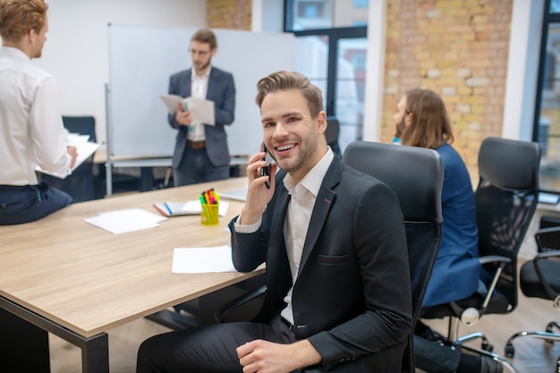 Alegre joven sonriente en traje de negocios hablando por teléfono inteligente en la mesa y colegas detrás de la oficina