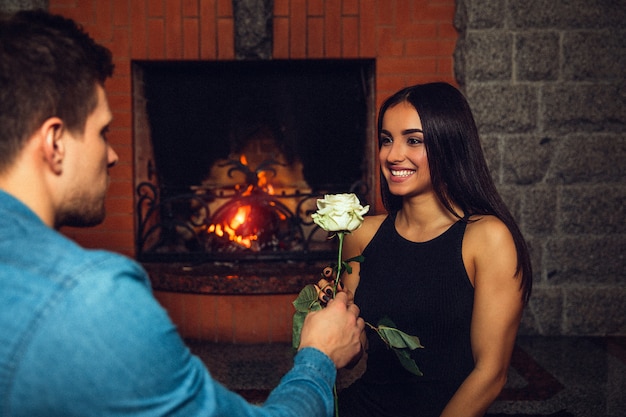 Alegre joven sonrisa a su novio. Ella lo mira. Guy dale una rosa blanca. Se sientan en la chimenea.