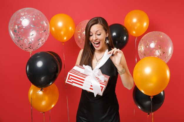 Alegre joven sorprendida en vestido negro celebrando, abriendo la caja roja con regalo presente en globos de aire de fondo rojo brillante. Día de San Valentín, feliz año nuevo, concepto de fiesta de vacaciones de maqueta de cumpleaños.