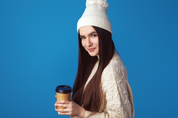 Alegre joven vestida de suéter blanco y sombrero sosteniendo café para llevar