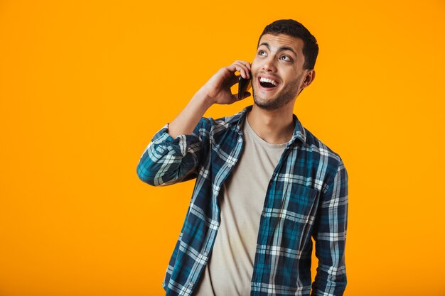 Foto alegre joven vistiendo camisa a cuadros se encuentran aisladas sobre pared naranja, hablando por teléfono móvil