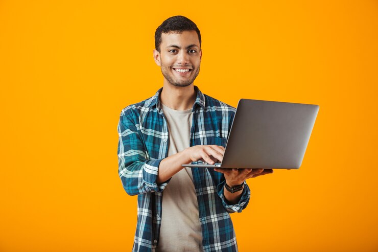 Foto alegre joven vistiendo camisa a cuadros que se encuentran aisladas sobre fondo naranja, utilizando equipo portátil