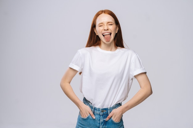 Alegre joven vistiendo camiseta y pantalones de mezclilla mostrando la lengua sobre fondo blanco aislado