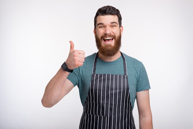 Alegre joven vistiendo un delantal de cocina o cocina está mostrando como gesto de aprobación o pulgar arriba