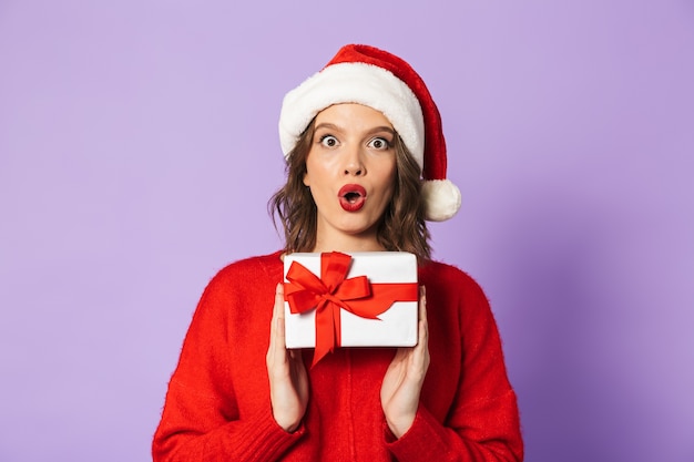 Alegre joven vistiendo rojo sombrero de Navidad que se encuentran aisladas sobre pared violeta, sosteniendo una caja de regalo