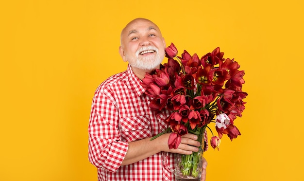 un alegre jubilado maduro con barba sostiene flores de tulipanes de primavera sobre fondo amarillo.