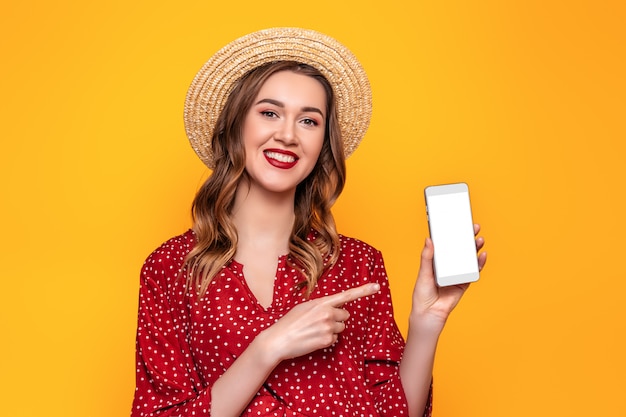 Alegre linda joven apuntando con un dedo en la pantalla blanca de un teléfono móvil aislada en un espacio de maqueta de pared amarilla para el diseño