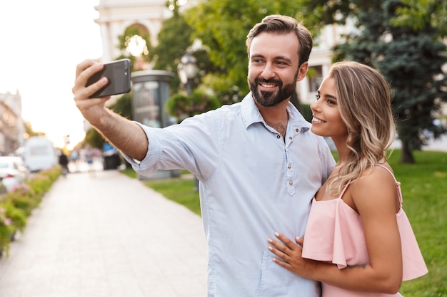 Alegre linda joven pareja amorosa caminando por la calle al aire libre tomar un selfie por teléfono móvil.