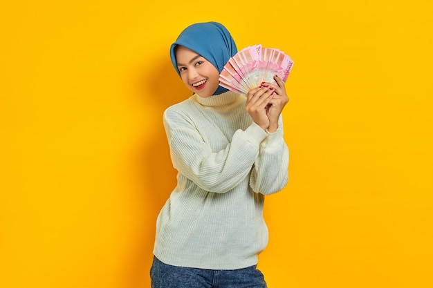 Alegre linda mulher muçulmana asiática em suéter branco mostrando fã de dinheiro em notas de rupia isoladas em fundo amarelo Conceito de estilo de vida religioso de pessoas