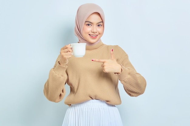 Alegre linda mulher muçulmana asiática no suéter marrom, apontando o dedo para a caneca de café isolada sobre fundo branco Conceito de estilo de vida religioso de pessoas