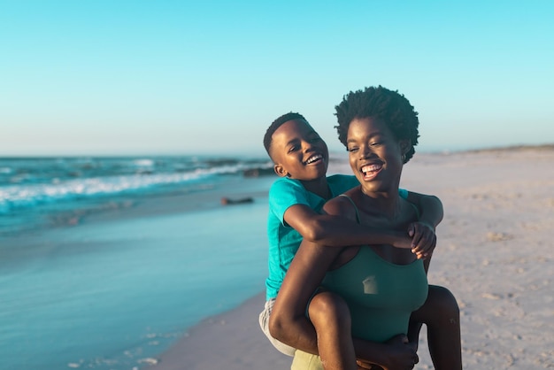 Alegre mãe afro-americana pegando carona no filho na praia contra o céu azul durante o pôr do sol. Copie o espaço, inalterado, família, juntos, infância, natureza, férias, diversão e conceito de verão.