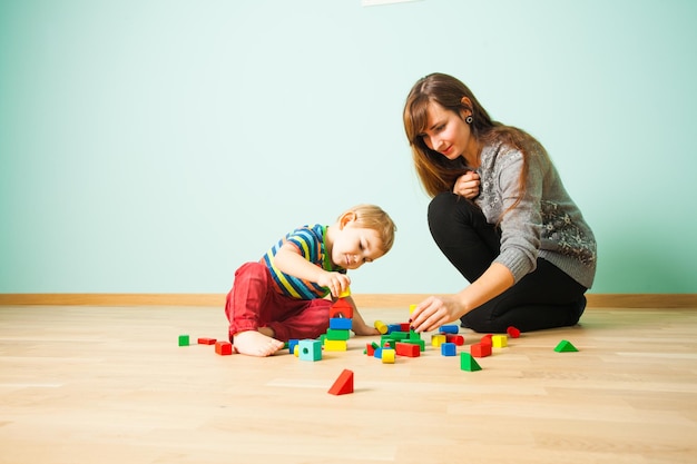 Alegre mãe com cabelo comprido brincando com seu filho em um piso de madeira no quarto. o menino está construindo torre de blocos de madeira multicoloridos.