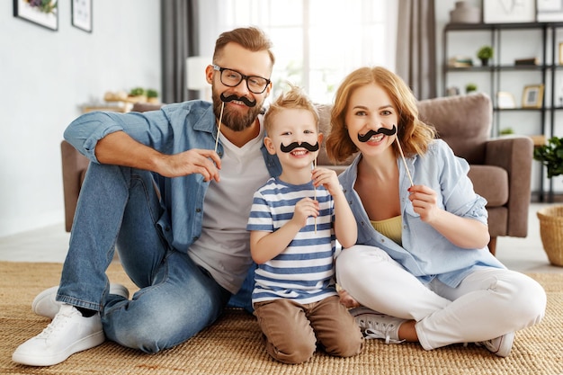 Alegre mãe e pai e menino bonito com bigode falso sorrindo e olhando para a câmera enquanto está sentado no chão perto do sofá na aconchegante sala de estar