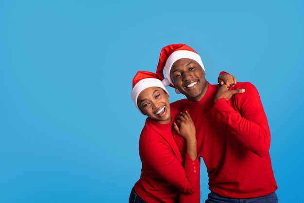 Alegre marido y mujer africanos con sombreros de Papá Noel abrazando el estudio