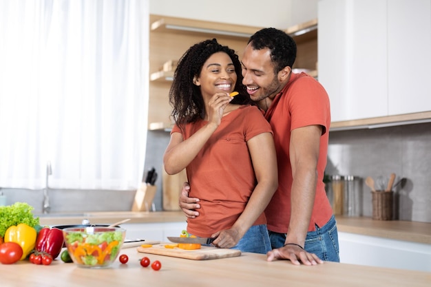 Alegre marido negro milenario abrazando a la mujer cortando verduras orgánicas en la ensalada y alimentando al hombre