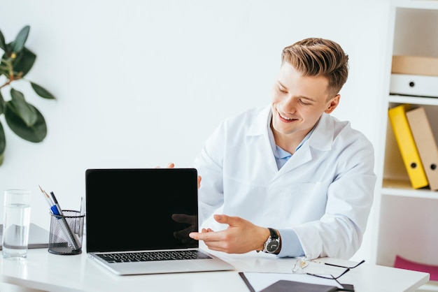 Alegre médico apontando com o dedo para laptop com tela em branco