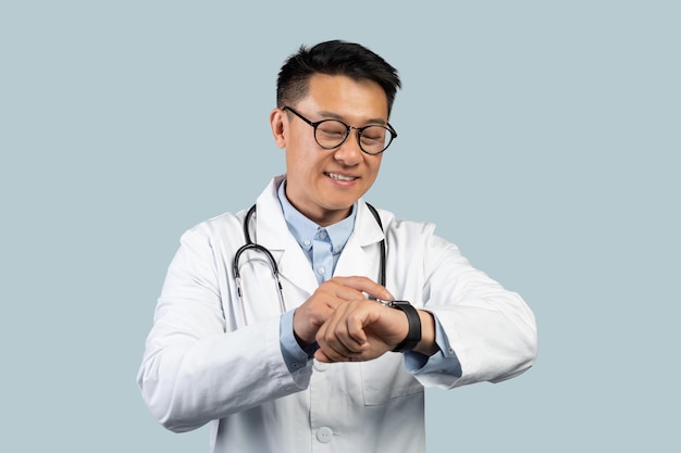 Un alegre médico chino de mediana edad con gafas de bata blanca mira el reloj a mano aislado de fondo azul