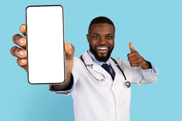 Alegre médico negro demonstrando smartphone em branco e mostrando o polegar para a câmera