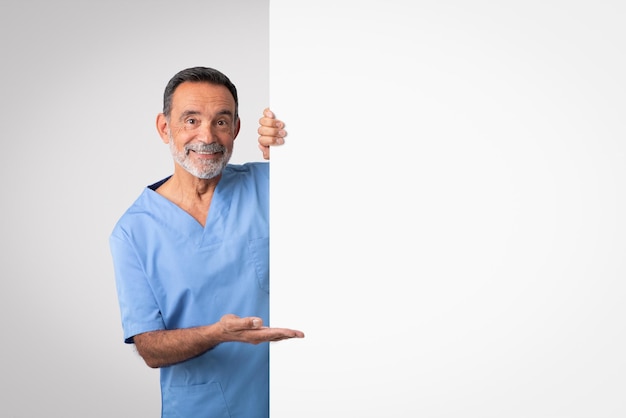 Alegre médico senior europeo en uniforme apunta a una gran pancarta con espacio libre en gris