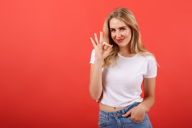 Alegre menina bonita jovem bonita bonita com cabelo loiro em t-shirt mostra gesto bem sobre fundo vermelho