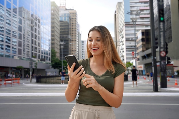 Alegre menina brasileira assistindo vídeo engraçado no telefone parado na rua durante o dia Cabelos castanhos com sorriso de dentes usa camiseta verde Conceito de vício em mídia social