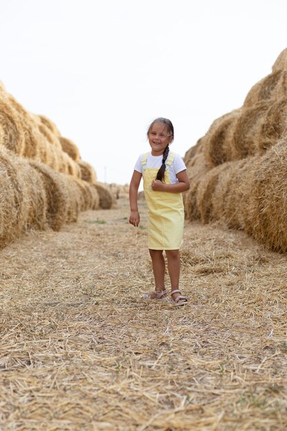 Alegre menina brincalhão com trança andando na estrada de campo de feno entre pilhas de feno olhando para a câmera vestindo vestido de verão se divertindo longe da cidade no campo cheio de feno dourado