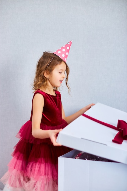 Alegre menina de vestido rosa e chapéu abre grande caixa de presente com balões em serpentinas de festa de aniversário em casa, feliz aniversário. A comemorar