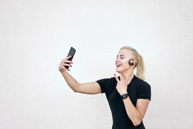 Alegre menina desportiva loira sorrindo suavemente vestindo roupas esportivas pretas, ouvindo música, tomando selfi e mostrando a vitória de cantar.