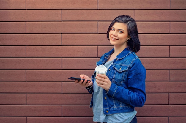 Alegre menina morena caucasiana com uma jaqueta jeans com smartphone em frente a parede de tijolos, segurando na mão café quente.