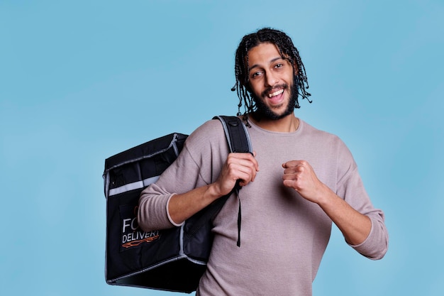 Alegre mensajero del servicio de entrega de alimentos de pie con mochila térmica y retrato sonriente. Joven repartidor despreocupado entregando comida para llevar en una bolsa y mirando a la cámara