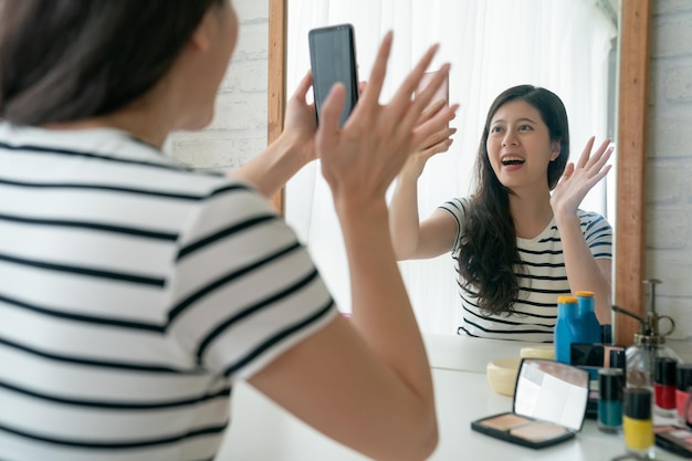 Alegre modelo feminino fazendo maquiagem na penteadeira no quarto. mulher asiática falando conversando em videochamada com fãs e público tutorial de gravação on-line aplicando o início cosmético da mão acenando.