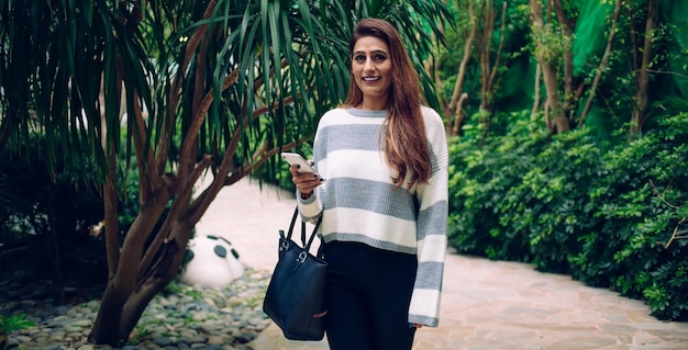 Alegre mujer adulta bastante sonriente en ropa casual con bolso paseando con teléfono inteligente