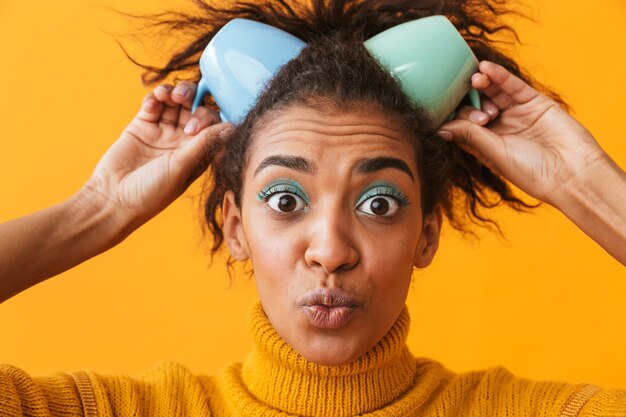 Alegre mujer africana vistiendo suéter sosteniendo tazas sobre su cabeza aislada