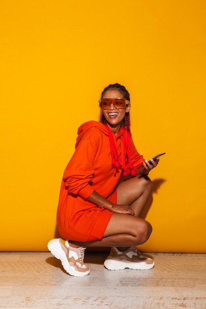 Alegre mujer afroamericana con gafas de sol y una sudadera con capucha sonriendo mientras sostiene el teléfono celular aislado sobre amarillo