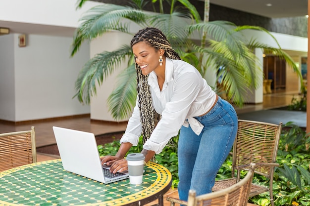 Alegre mujer afroamericana de pie usando la computadora portátil en una tienda de café afuera. Mujer con trenzas usando laptop y tomando café.