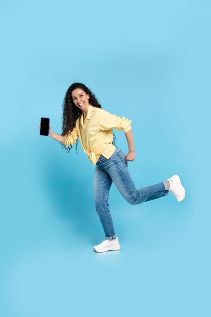 Alegre mujer árabe mostrando la pantalla en blanco del teléfono sobre fondo azul.