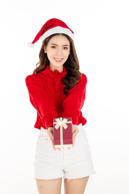 Alegre mujer asiática atractiva con un vestido rojo con un gorro de Papá Noel sostiene una caja de regalo sobre fondo blanco aislado. Bella dama con cabello largo y negro.