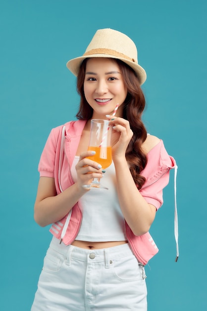 Alegre mujer asiática bebiendo jugo de frutas y vistiendo sombrero de paja, camiseta blanca, jeans cortos, chaqueta rosa sobre fondo azul.