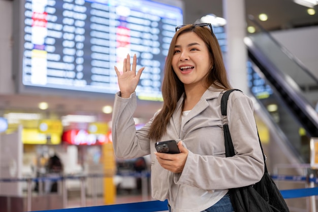 Una alegre mujer asiática está agitando la mano y llamando a sus amigos mientras está de pie en el aeropuerto