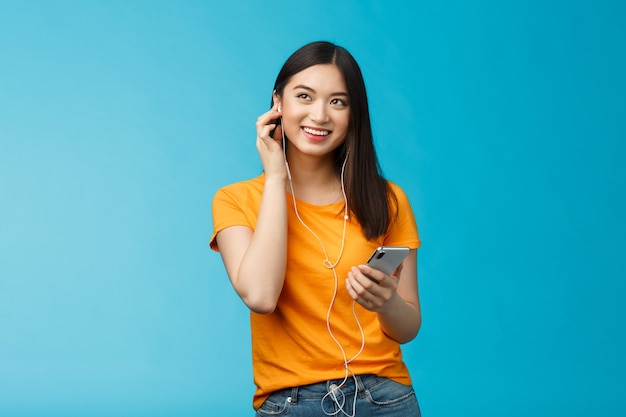 Alegre mujer asiática guapa mirar hacia arriba encantada, sostener el auricular enchufable del teléfono inteligente sonriendo satisfecho con buena calidad de sonido, disfrutar escuchando música atril fondo azul entusiasta optimista