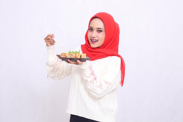 Alegre mujer asiática en hijab sosteniendo palillos y levantando un plato que contiene sushi japonés f