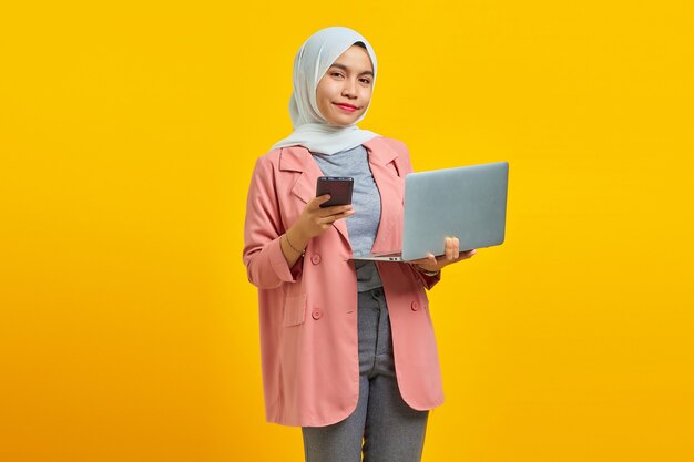 Alegre mujer asiática joven sonriente sosteniendo un teléfono móvil y un portátil sobre fondo azul.