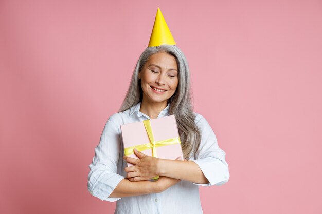 Alegre mujer asiática madura con cabello gris suelto abraza la caja de regalo posando en el estudio