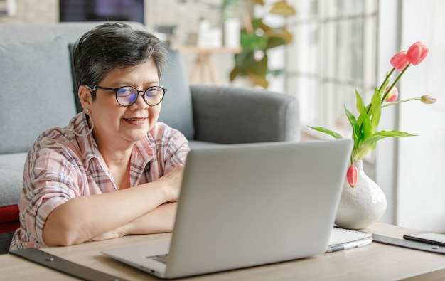 Alegre mujer asiática madura gesticulando y hablando con un colega en línea mientras está sentado cerca del sofá y trabaja de forma remota desde casa durante el día