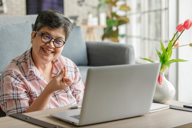 Alegre mujer asiática madura gesticulando y hablando con un colega en línea mientras está sentado cerca del sofá y trabaja de forma remota desde casa durante el día