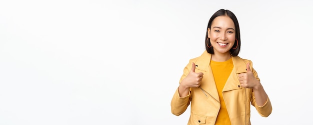 Alegre mujer asiática sonriendo complacida mostrando los pulgares hacia arriba en aprobación de pie en ropa informal sobre fondo blanco.