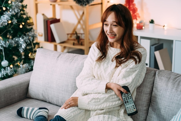 una alegre mujer asiática sostiene su vientre y su controlador de televisión mientras disfruta de divertidos espectáculos con una risa durante las vacaciones de invierno de Navidad en el sofá de la sala de estar en casa